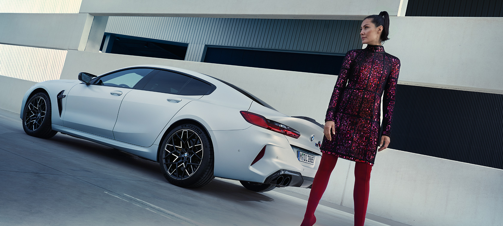 O BMW M8 Competition Gran Coupé, F93, atualização, 2022, pintura branco Frozen Brilliant BMW Individual metalizada, perspetiva traseira a três quartos com um modelo feminino parado ao lado do veículo